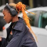 A person with an orange braided bun and ponytail, wearing black headphones and a black cropped jacket, looks at their phone.