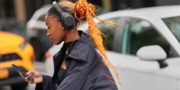 A person with an orange braided bun and ponytail, wearing black headphones and a black cropped jacket, looks at their phone.