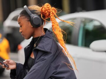 A person with an orange braided bun and ponytail, wearing black headphones and a black cropped jacket, looks at their phone.