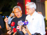 Two men at a press conference surrounded by microphones, one speaking, another listening attentively.