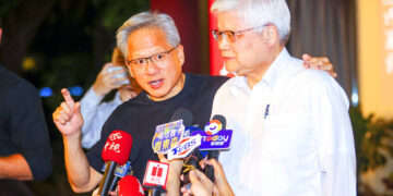 Two men at a press conference surrounded by microphones, one speaking, another listening attentively.
